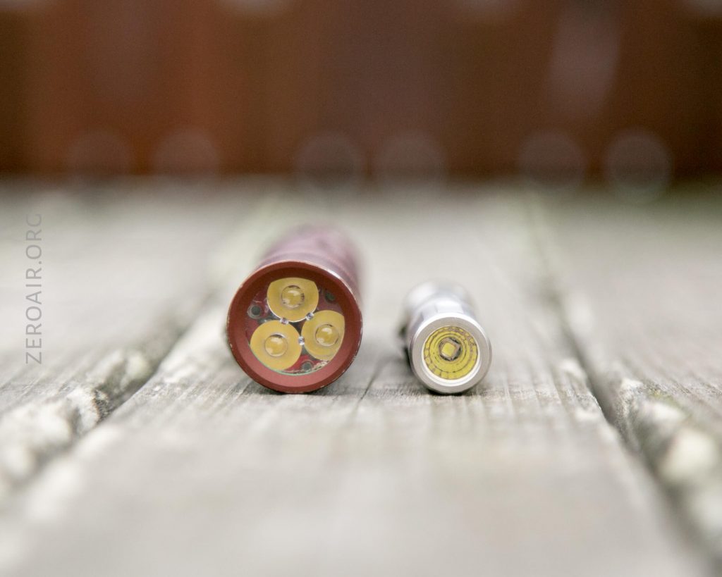 A close-up of two batteries standing on a wooden surface; the left battery is larger and brown with three contacts, while the right battery is smaller and silver with a single contact.