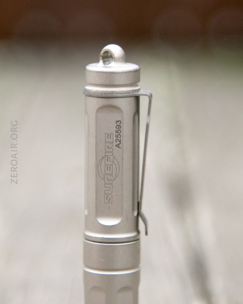 A close-up of a silver SureFire flashlight with a pocket clip, serial number A25693, and the SureFire logo engraved on the side; the background is blurred.