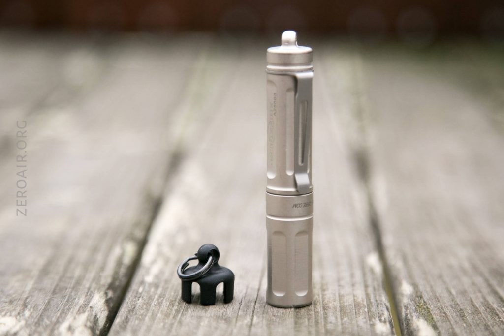 A small metallic flashlight stands upright on a wooden surface next to a black keychain accessory. The background is blurred, and the flashlight features a sleek, cylindrical design.