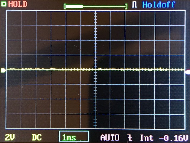 An oscilloscope screen displays a mostly flat yellow waveform along the center, with settings and indicators such as HOLD, 2V DC, and 1ms visible along the edges.
