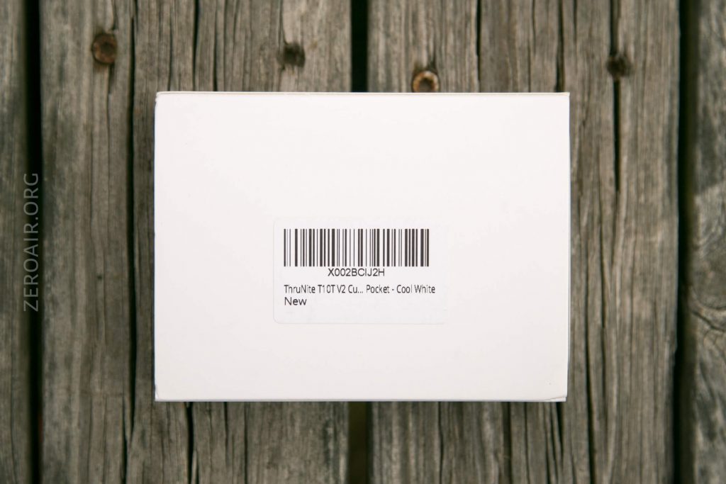 A white rectangular box with a barcode label on top, placed on a weathered wooden surface. The label reads ThruNite TCTV2 Cu. Pocket - Cool White New. The word ZEROAIR.ORG is on the left side.