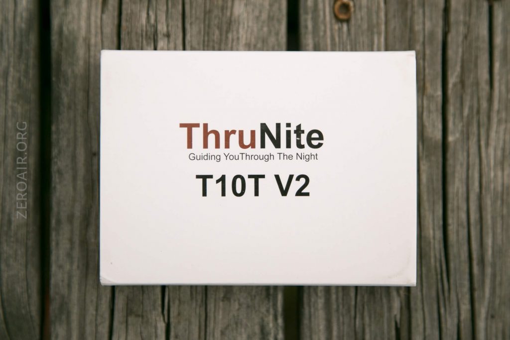 A white box labeled ThruNite Guiding You Through The Night T10T V2 rests on a wooden surface. The brand name is in brown and black text, with the product name in bold black letters.