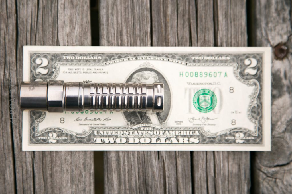 A metal object with grooves is placed on top of a two-dollar bill, both resting on a weathered wooden surface.