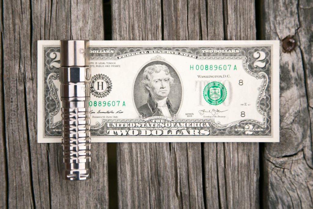 A metal pen with a grooved design rests horizontally on top of a two-dollar bill, which is placed on a wooden plank surface with visible grain and gaps.