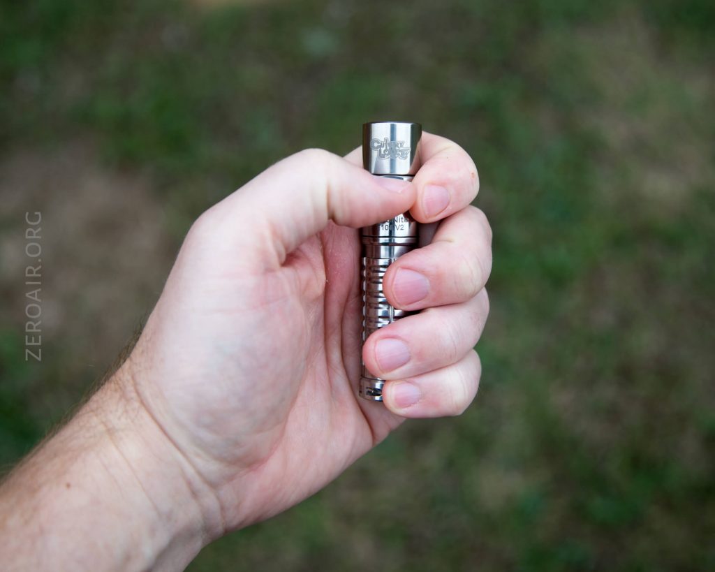 A hand holding a small, metallic, cylindrical flashlight outdoors with green grass in the blurred background. The hand is gripping the flashlight firmly, with fingers wrapped around its body.
