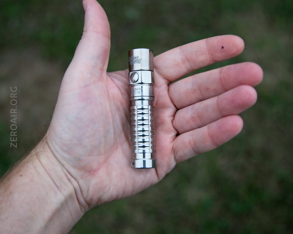 A persons left hand holding a small, metallic, cylindrical flashlight with a ribbed grip, against a blurred outdoor background. The word ZEROAIR.ORG is visible along the left side of the image.