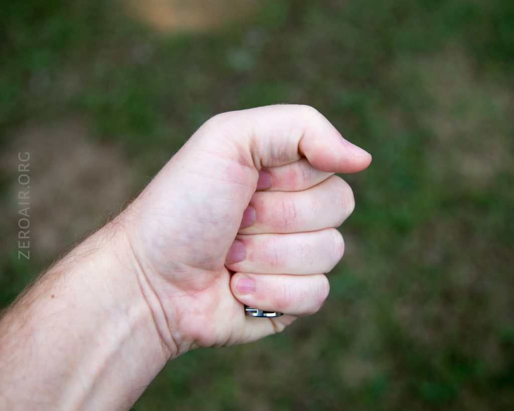 A close-up of a left hand forming a fist with the thumb tucked over the fingers, held in front of a blurred outdoor background with grass and dirt visible. The word ZEROAIR.ORG is faintly visible on the left side.