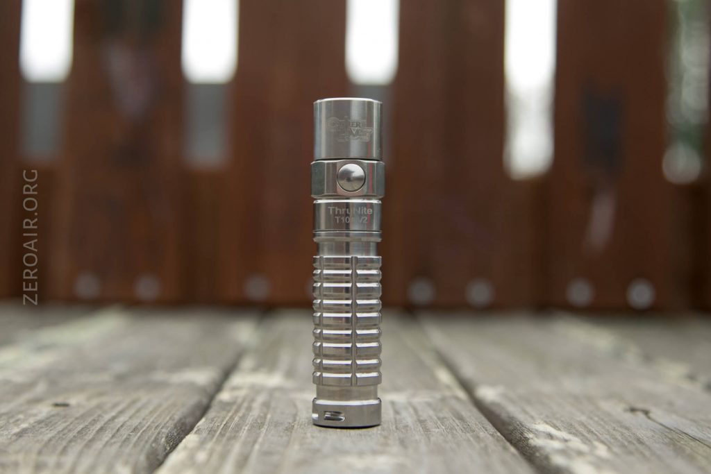 A stainless steel Thrunite T1T2 flashlight stands upright on a wooden surface with a blurred wooden fence in the background. The flashlight has a grooved cylindrical body and branding visible on the head.