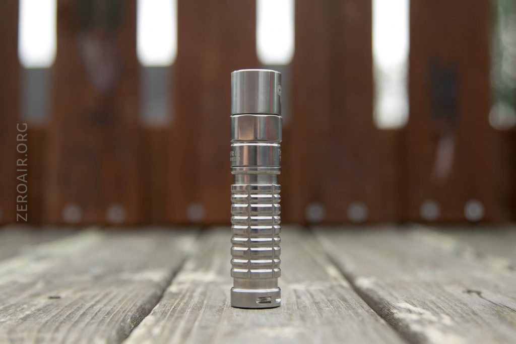 A metallic, cylindrical flashlight with a ribbed grip stands upright on a wooden surface, with a blurred brown fence in the background. The word ZEROAIR.ORG is faintly visible on the left side of the image.