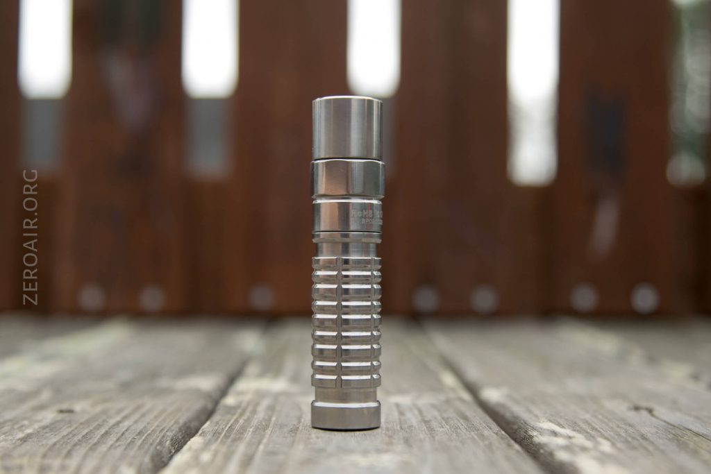 A stainless steel flashlight stands upright on a wooden surface with a blurred wooden fence in the background. The flashlight has a ribbed grip and a cylindrical shape.