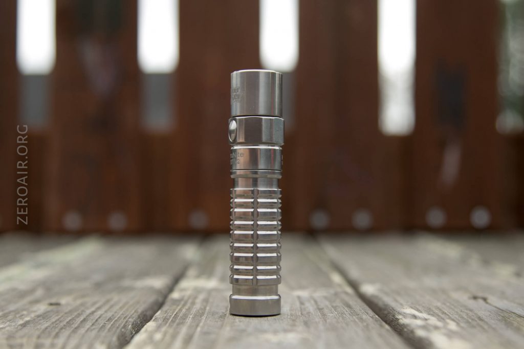 A small, metallic flashlight with a ribbed grip stands upright on a wooden surface. The background is blurred, showing vertical brown slats and some light coming through gaps.