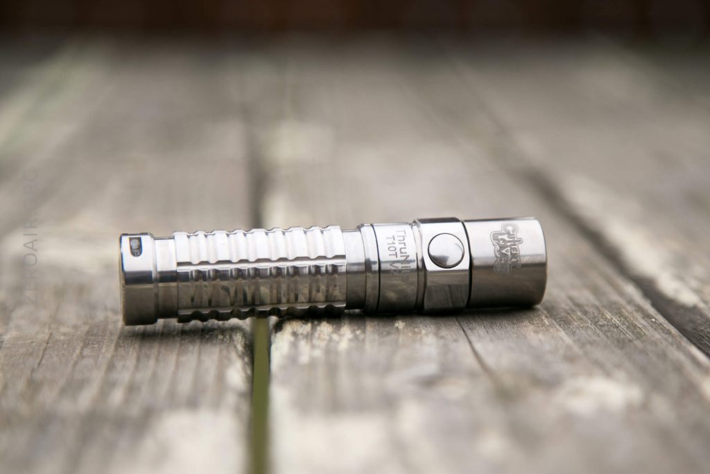 A metallic flashlight with a ribbed grip lies on its side on a weathered wooden surface, with engraved text and symbols visible on its body.