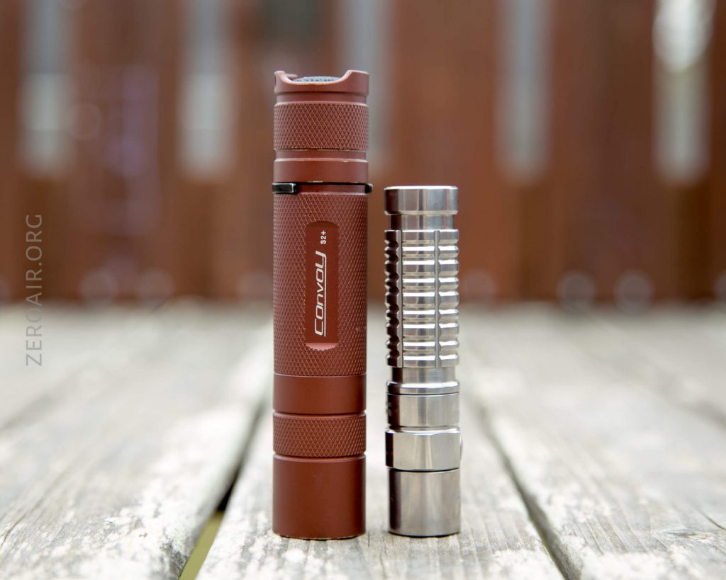 A brown Convoy S3 flashlight stands upright next to a shorter, silver metallic flashlight on a wooden surface. The background is out of focus, with ZERGAIR.ORG faintly visible on the left side.