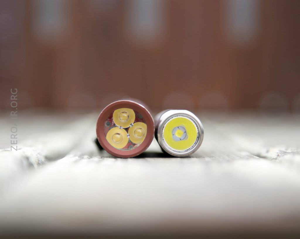 Two flashlight heads are shown from the front, placed side by side on a wooden surface. The left flashlight has three yellow LED emitters, and the right flashlight has a single yellow LED emitter.