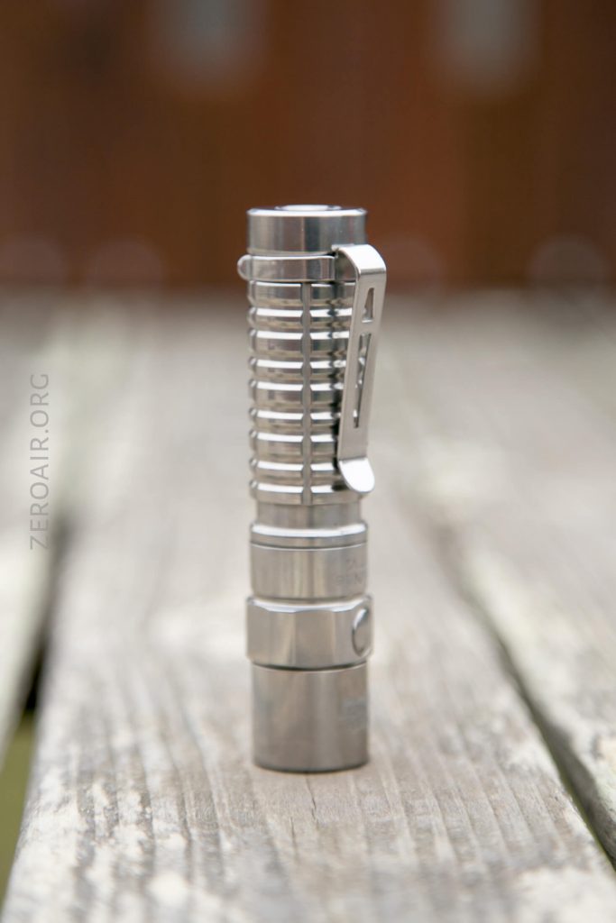 A metallic flashlight with a ridged design and a pocket clip stands upright on a weathered wooden surface.