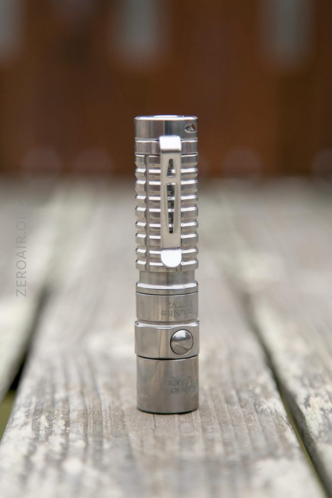 A stainless steel flashlight with a ribbed grip and clip stands upright on a weathered wooden surface, with a blurred brown background.