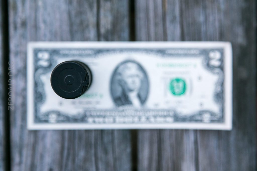 A black cylindrical object is positioned vertically above the center of a two-dollar bill, which is placed on a wooden surface. The image is taken from above, with the bill slightly out of focus.