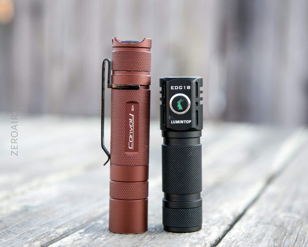 Two compact flashlights stand upright on a wooden surface. The left flashlight is maroon with a clip and labeled Convoy S2+. The right flashlight is black and labeled Lumintop EDC18. The background is blurred.