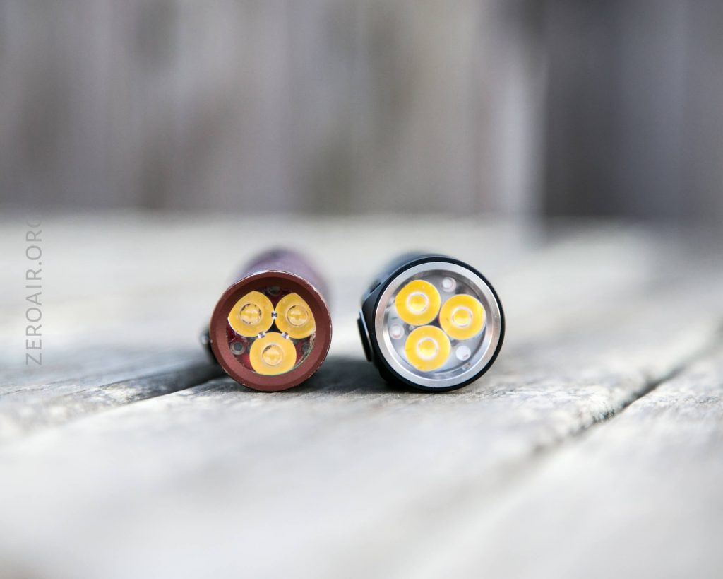 Two compact cylindrical flashlights lie side by side on a wooden surface, viewed from the front to show three LED bulbs arranged in a triangle pattern in each device.