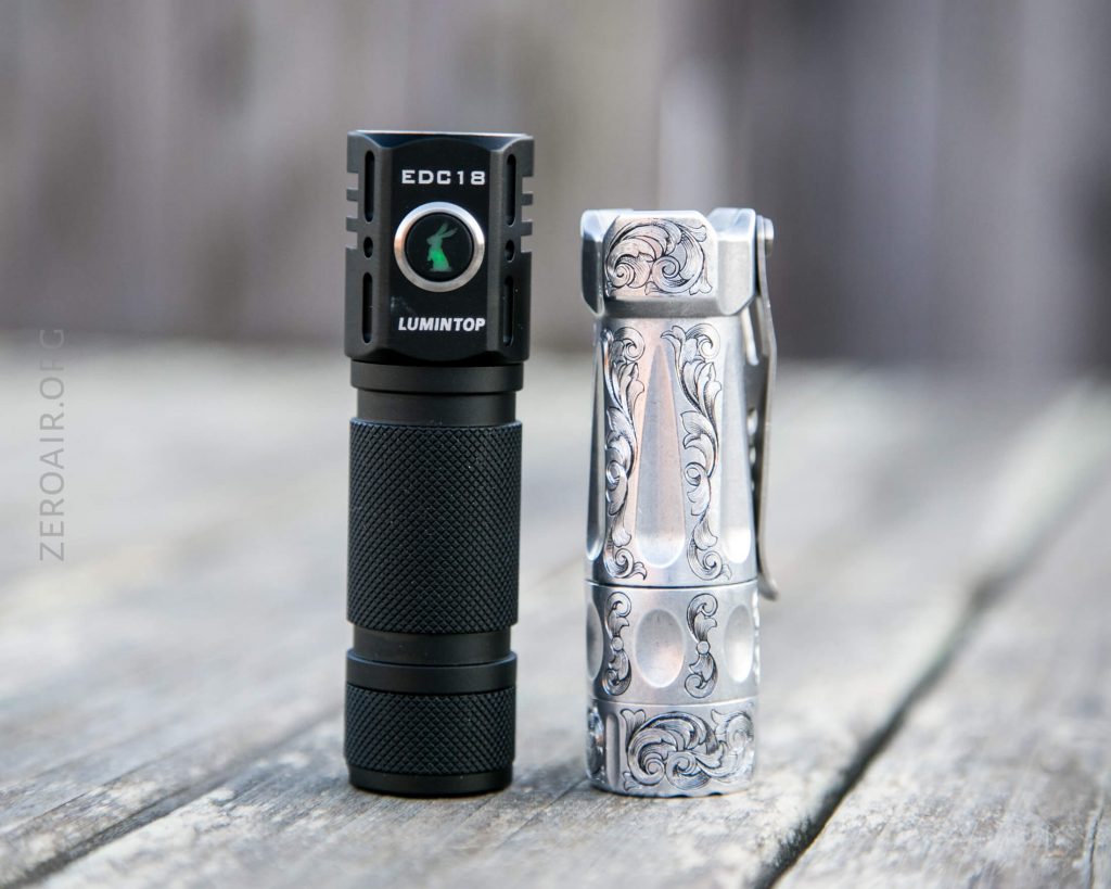 Two flashlights stand upright on a wooden surface. The left flashlight is black and labeled EDC18 LUMINTOP, while the right one is silver with ornate, engraved designs and a metal pocket clip.