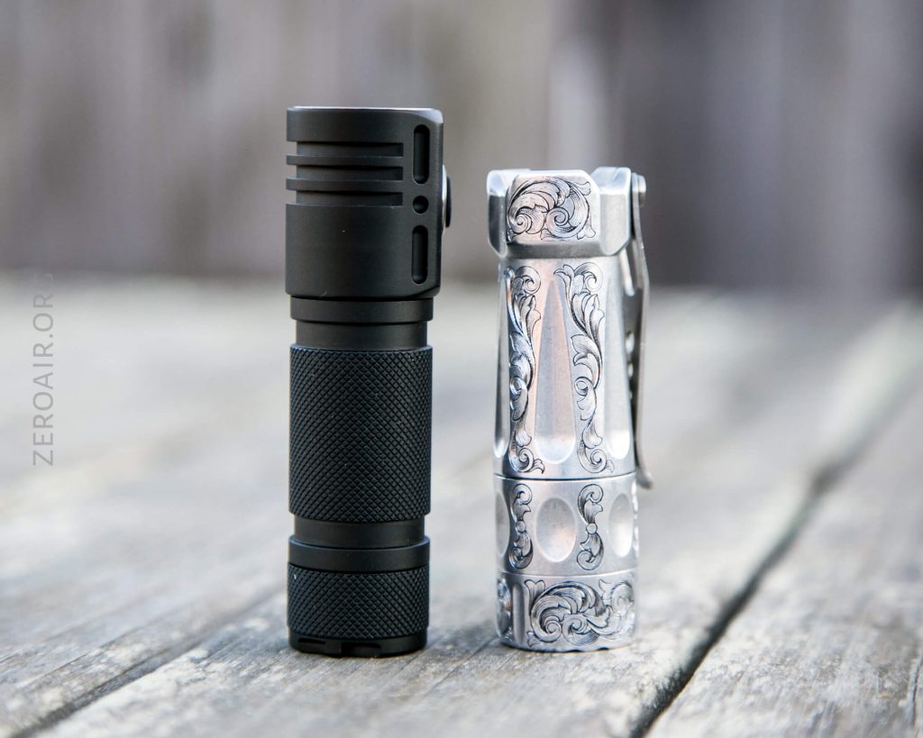 Two compact flashlights are standing upright on a weathered wooden surface. The flashlight on the left is black with a textured grip, while the one on the right is silver with ornate, engraved patterns.