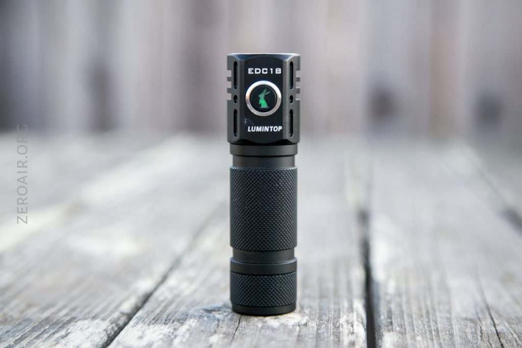 A black LUMINTOP EDC18 flashlight stands upright on a weathered wooden surface with a blurred wooden background. The flashlight has a textured grip and branding visible on its body.