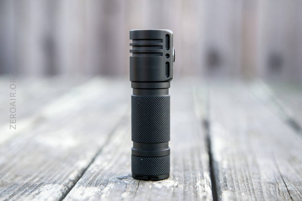 A small black flashlight standing upright on a weathered wooden surface, with a blurred wooden background. The flashlight has a textured grip and vent-like grooves near the top.