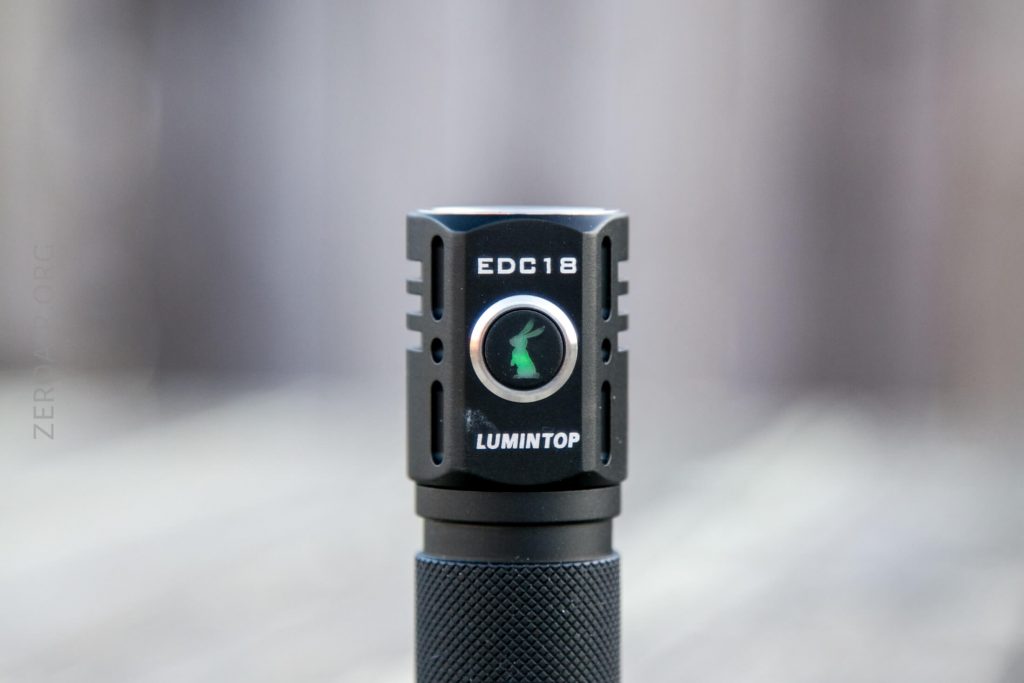 A close-up of a black Lumintop EDC18 flashlight, showing its square head with visible button featuring a green rabbit logo and engraved text EDC18 above LUMINTOP. The background is blurred.
