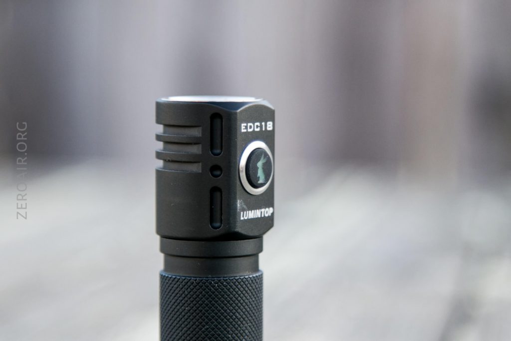 A close-up of a black LUMINTOP EDC18 flashlight standing upright on a wooden surface, with EDC18 and LUMINTOP labels visible on the head. The background is blurred.