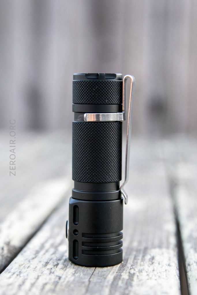 A small black flashlight with a textured grip and metal pocket clip stands upright on a weathered wooden surface, with a blurred background.