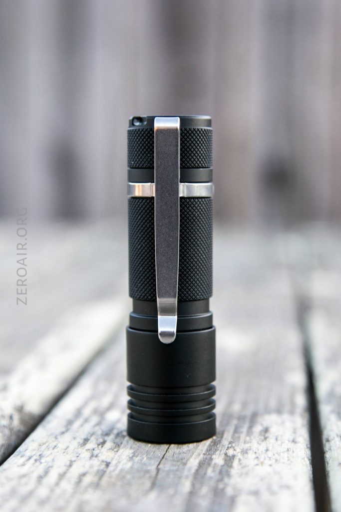 A small black flashlight with a textured grip and metal clip stands upright on a weathered wooden surface, with a blurred grey and brown background.