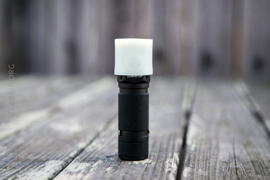 A black flashlight with a white diffuser cap stands upright on a weathered wooden surface. The background is blurred, featuring vertical wooden planks.