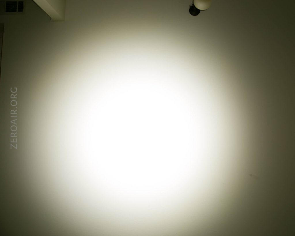 A bright white circular light is projected onto a wall in a dim room. A ceiling corner and a light fixture are visible at the top right. The text ZEROAIR.ORG appears faintly along the left side.
