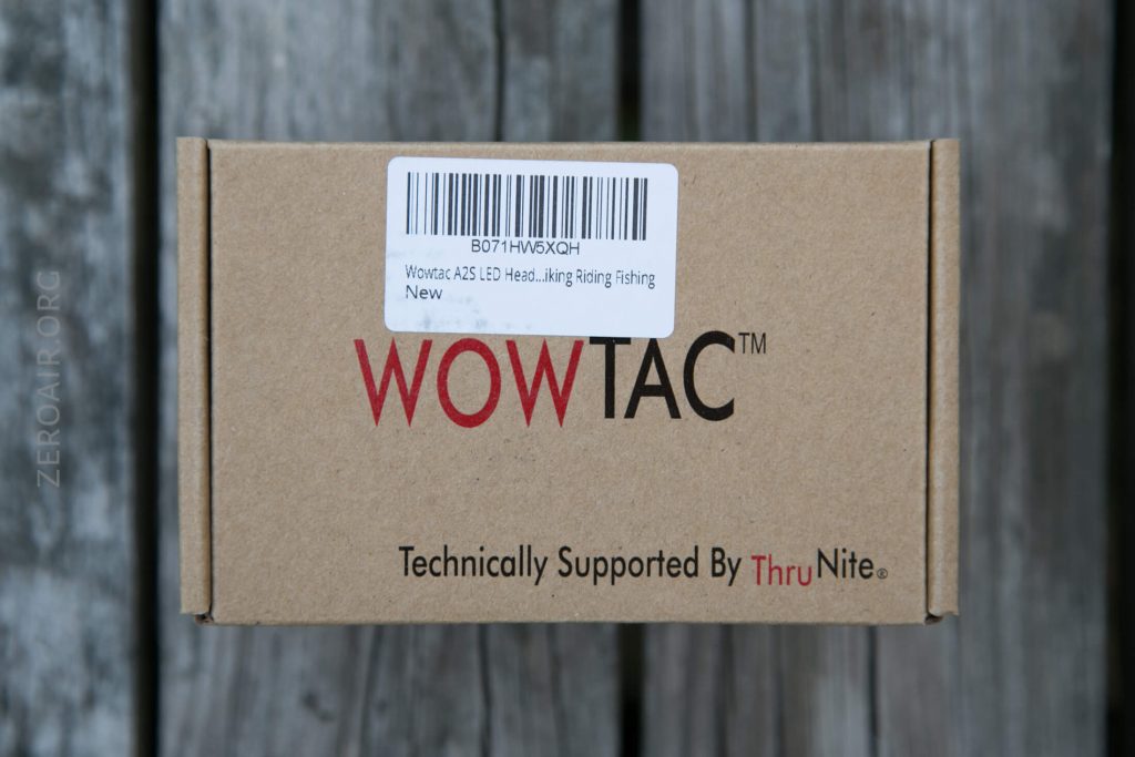 A cardboard box with the brand name WOWTAC and the text Technically Supported By ThruNite printed on it. A shipping label on top mentions Wovtac A25 LED Head…king Riding Fishing New. The box is on a wooden surface.