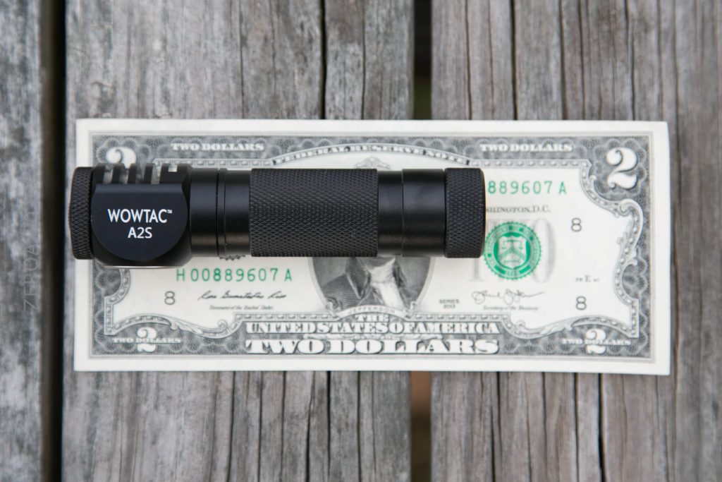 A black WOWTAC A2S flashlight is placed horizontally on a two-dollar bill, both resting on a weathered wooden surface.