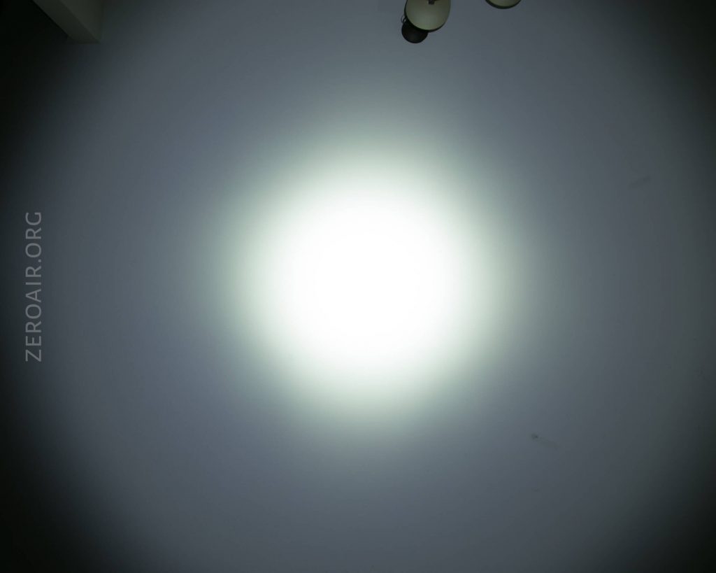 A bright, circular white light is projected onto a smooth gray wall. The text ZEROAIR.ORG appears vertically along the left side. Three round light fixtures are partially visible in the upper right corner.
