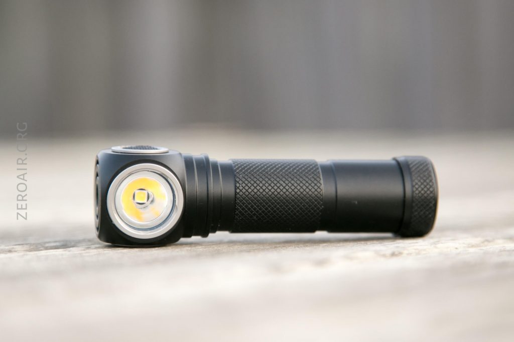 A small black flashlight with a right-angle head and textured grip lies on a wooden surface. The lens and LED are visible on the left. The background is blurred.