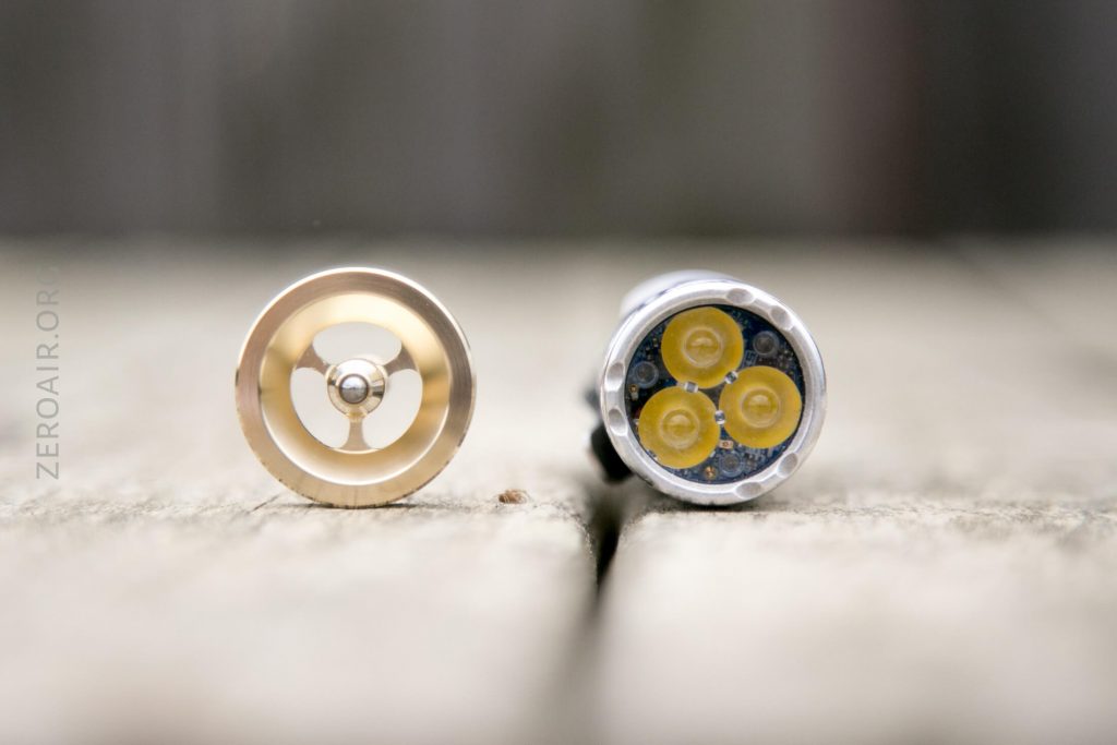 Close-up view of two cylindrical objects standing upright on a wooden surface; the left object shows a metal ring with a central sphere, while the right object reveals three yellow circular components inside.