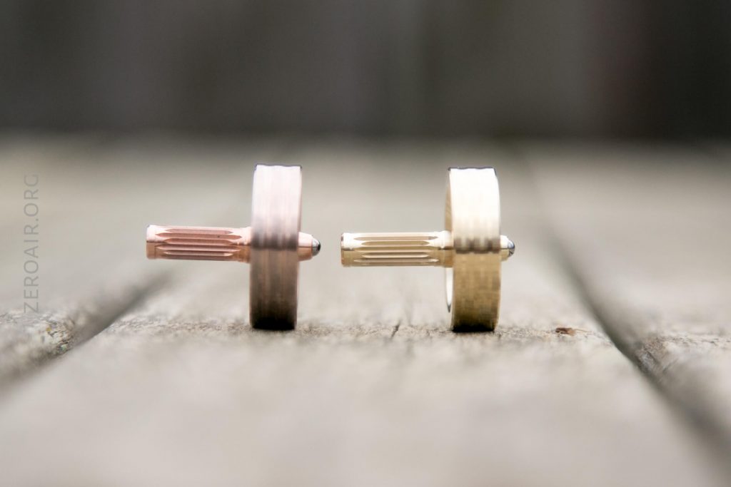 Two metal mechanical pencil tips, one rose gold and one gold, positioned horizontally and facing each other on a wooden surface, with the background blurred.