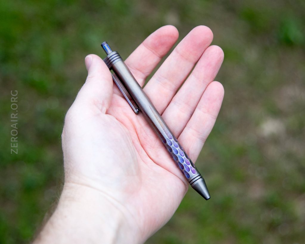 A person holds a metallic pen with a textured, hexagonal grip pattern in their open palm. The background is blurred green grass. The word ZEROAIR.ORG is visible along the left side.
