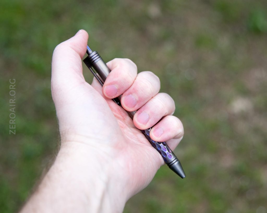 A right hand holding a metallic pen with a purple pattern, positioned outdoors with a blurry green background. The pen is held between the thumb and fingers, point facing outward.
