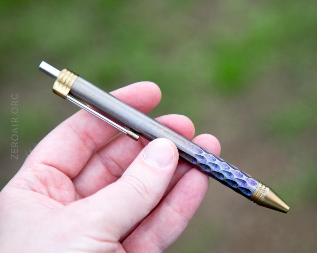 A hand holds a metallic pen with gold-colored accents and a textured, multicolored grip area against a blurred green background.