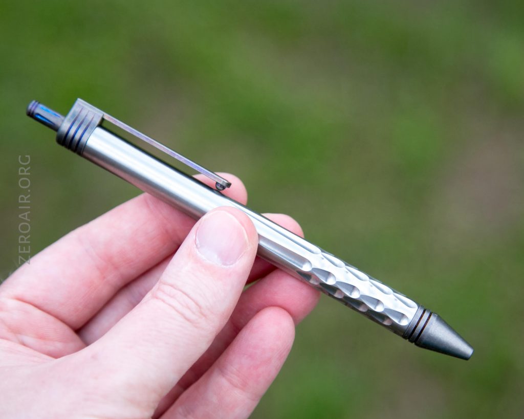 A hand holding a metallic pen with a textured, dimpled grip area, a black clip, and a pointed tip. The background is blurred green.