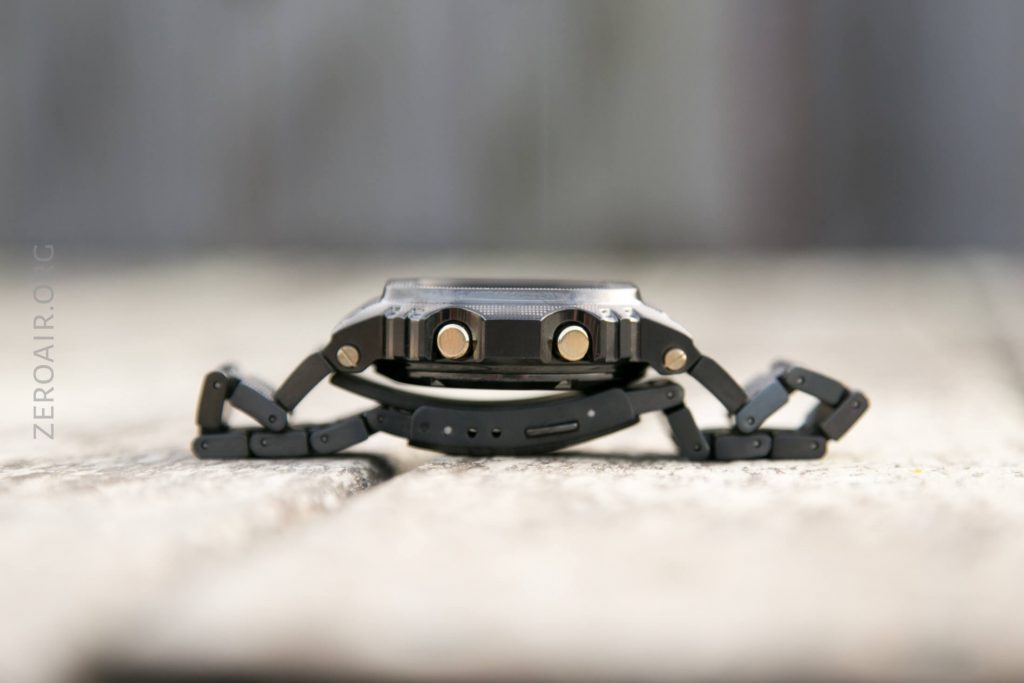 A close-up side view of a black wristwatch with a segmented band and two prominent buttons, placed on a wooden surface. The background is blurred.