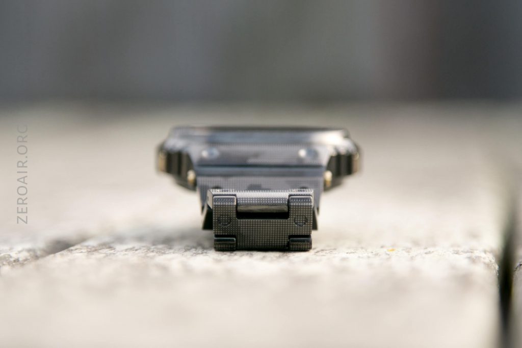A black, rectangular digital watch lies face up on a wooden surface, photographed from the front at a low angle. The focus is on the watch’s clasp, with the rest of the watch and background blurred.