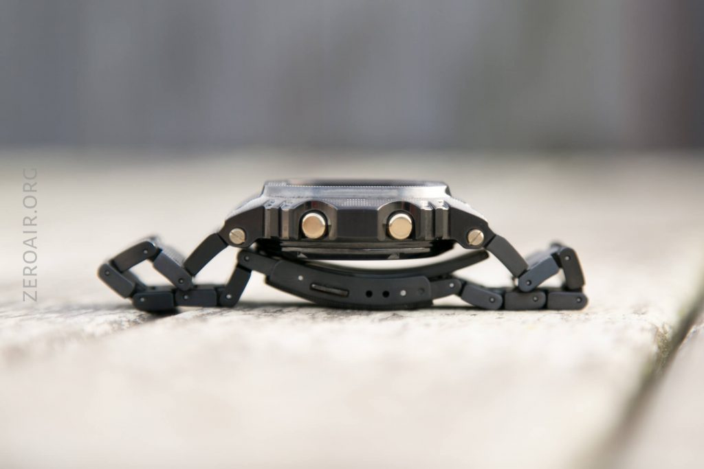 A close-up side view of a black wristwatch with a metal band laid flat on a light-colored wooden surface, showing two buttons on the watch case and the band partially unclasped.