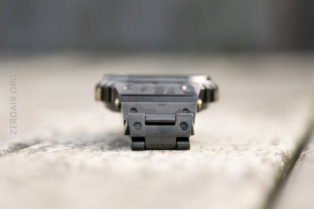 A close-up, front-facing view of a compact black flashlight with a textured surface, placed on a light wooden surface. The background is softly blurred. The text ZEROAIR.ORG is vertically visible on the left side.