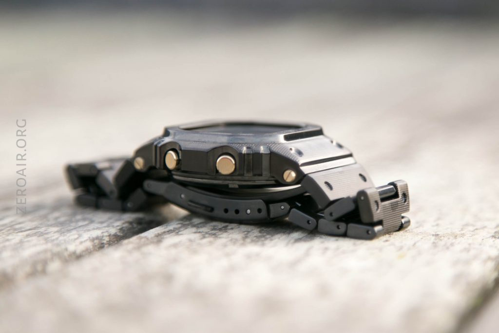 A close-up side view of a black digital wristwatch with metallic buttons, lying on a weathered wooden surface. The focus is on the watch, showing its thickness and strap detail.