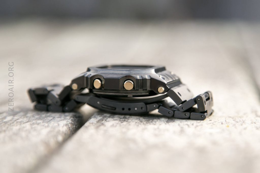 A close-up side view of a black digital wristwatch with a metal band lying flat on a light wooden surface. The image shows the watchs buttons and texturing in detail.