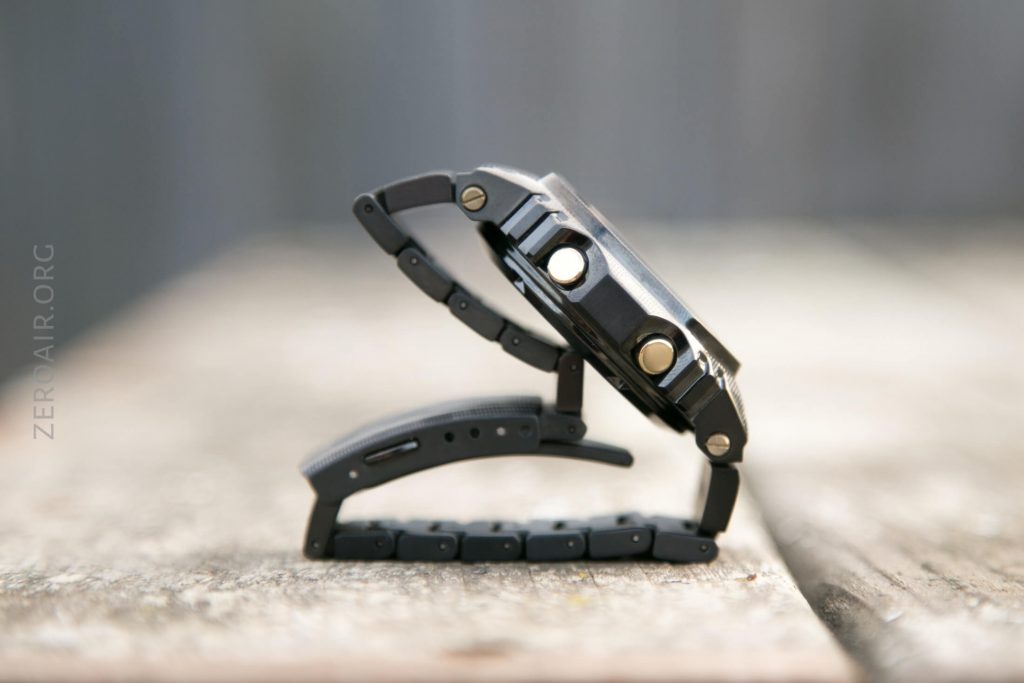 A black wristwatch with a metal band is standing upright on a wooden surface, positioned so the watch and band form a triangle. The background is blurred gray and brown tones.